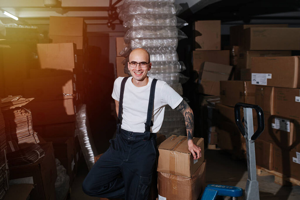 Charming male warehouse worker leaning on stack of carton boxes on pallet jack - Foto, afbeelding