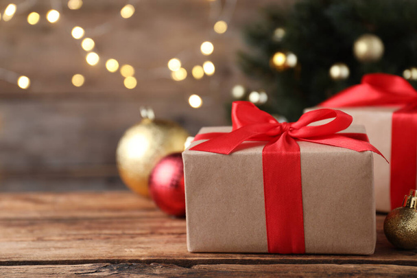 Christmas gift box with red ribbon and golden bauble on wooden table against blurred lights, space for text - Φωτογραφία, εικόνα