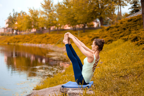 Slender young brunette yogi performs challenging yoga exercises on the grass in autumn against the background of nature. beautiful sporty girl sitting in Both big toe exercise, Paripurna Navasana pose. - Photo, Image