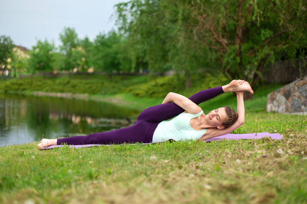 Slender young brunette yogi performs challenging yoga exercises on the green grass in the summer against the backdrop of nature. - Photo, Image