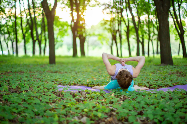 Thin brunette girl plays sports and performs yoga poses in a summer park on a sunset background. Woman doing exercises on the yoga mat. Summer forest. Soft focus. - Фото, изображение