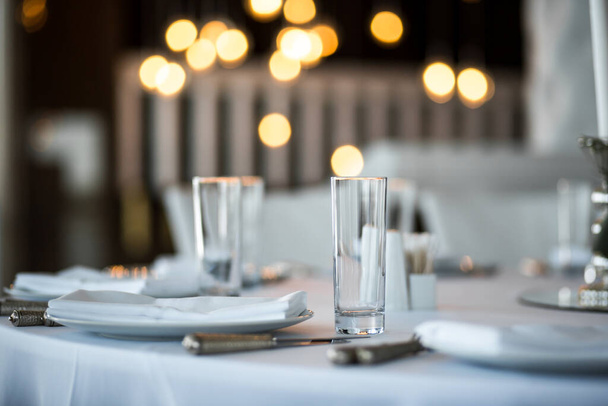 Table setting at a restaurant. Clean glass cup close up. Details of the banquet table. On the background blur are burning garlands with light bulbs. - Фото, изображение