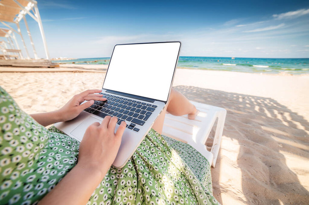 Mockup image of woman using laptop with blank white desktop screen while sitting on beach chair against blue sky background - Fotografie, Obrázek