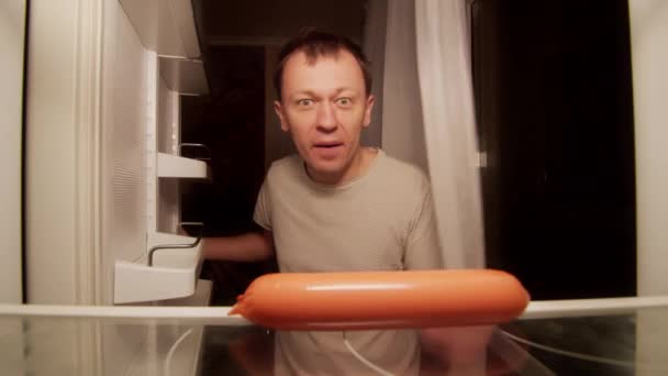 L'uomo affamato prende una salsiccia dallo scaffale del frigorifero, la morde - Filmati, video