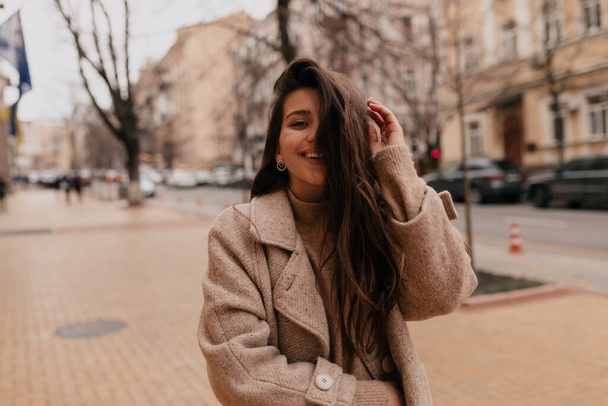 Stylish woman with long hair walking in the city wearing beige coat and posing with wonderful smile. High quality photo - Foto, Imagem