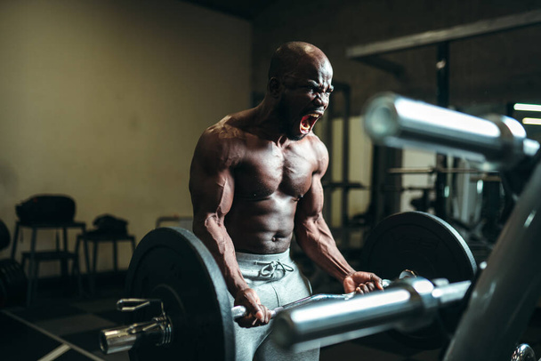 Emotional muscular african american man straining muscles, screaming and lifting a heavy barbell - Photo, image