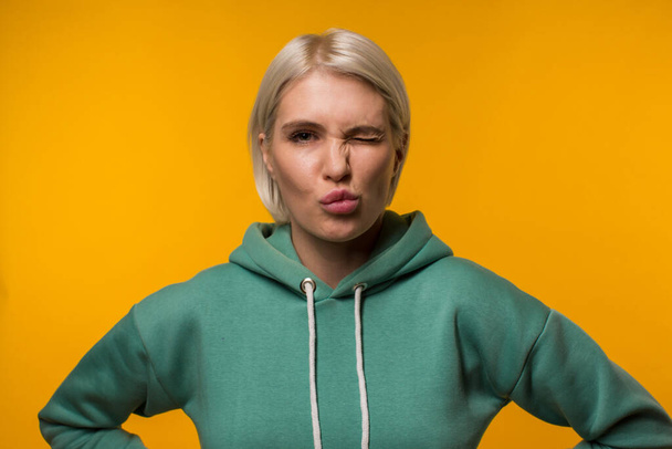 Blonde in green hoodie on a yellow background winks at the camera - Zdjęcie, obraz