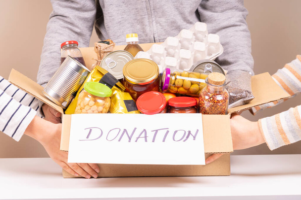 Voluntários mãos segurando caixa de doações de alimentos com produtos de supermercado na mesa branca - Foto, Imagem