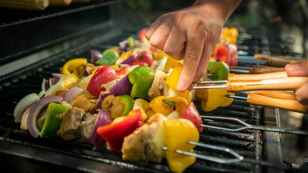 Hand of man with BBQ grill. Cook a delicious skewers on grilled meat. Food cooked with grilling barbecue. Chef cooking brochette in backyard of a house - 写真・画像