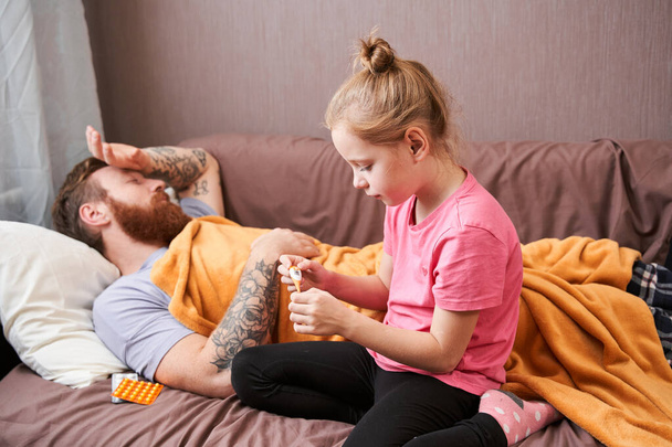 Man covered in blanket is lying on couch while his daughter is taking his temperature - Foto, Imagem