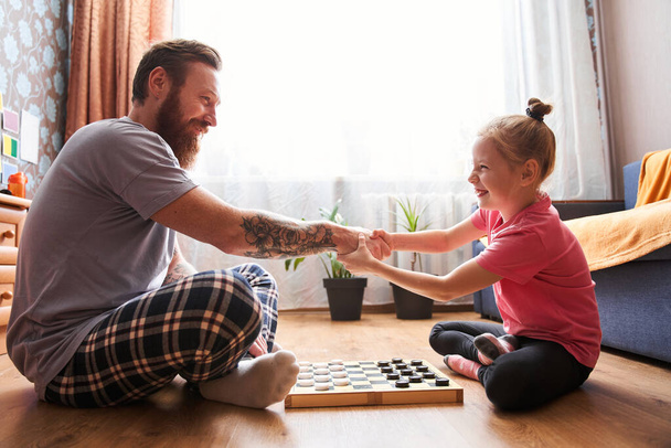 Girl and her handsome bearded dad in pajamas are smiling while playing checkers - Foto, Bild