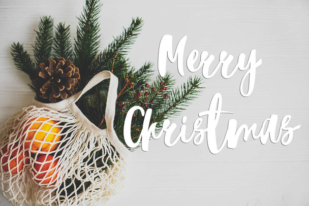 Frohe Weihnachten handgeschriebenen Text auf Baumwolltasche mit grünen Fichtenzweigen, Orangen und Tannenzapfen auf weißen rustikalen Tisch, keine Abfälle. Grußkarte, moderne Jahreszeiten. Frohe Feiertage - Foto, Bild