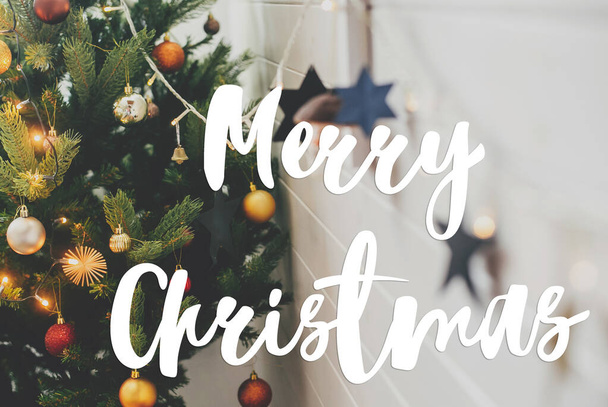 Καλά Χριστούγεννα κείμενο χειρόγραφο σε μοντέρνο διακοσμημένο χριστουγεννιάτικο δέντρο με κόκκινα και χρυσά στολίδια και εορταστικά φώτα. Ευχετήριες κάρτες, μοντέρνες εποχές. Καλές γιορτές. - Φωτογραφία, εικόνα