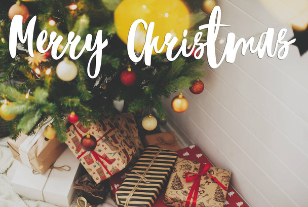 Noel ağacının altında, kırmızı ve altın renkli ışıklarla süslü Noel hediyeleri üzerine yazılmış mutlu Noeller mesajı. Tebrik kartı, modern mevsimler selamları. İyi tatiller. - Fotoğraf, Görsel