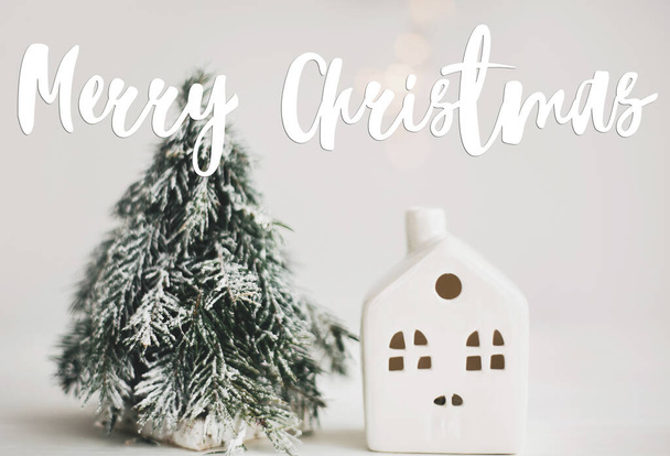 Joyeux texte de Noël écrit à la main sur la petite maison de Noël et l'arbre enneigé avec des lumières festives bokeh sur fond blanc, scène de vacances d'hiver. Carte de voeux, voeux des saisons modernes - Photo, image