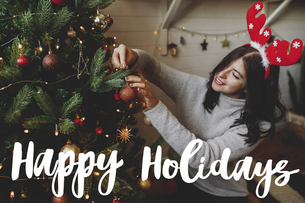 Boldog ünnepeket szöveg kézzel írt boldog nő rénszarvas agancs díszítő modern karácsonyfa csecsebecsével. Üdvözlőlap, modern évszakok köszöntése - Fotó, kép
