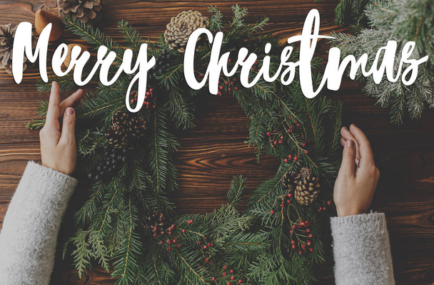 Wesołych Świąt tekst ręcznie napisany na rustykalnym wieńcu Bożego Narodzenia w rękach na drewnianym stole, leżał płasko. Kartka z życzeniami, współczesne pozdrowienia. Wesołych świąt - Zdjęcie, obraz