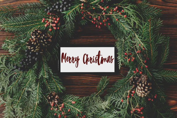 Merry Christmas text handwritten on phone screen in rustic christmas wreath on wooden table flat lay, online greetings. Greeting card, modern seasons greetings. Happy holidays - Foto, Imagem