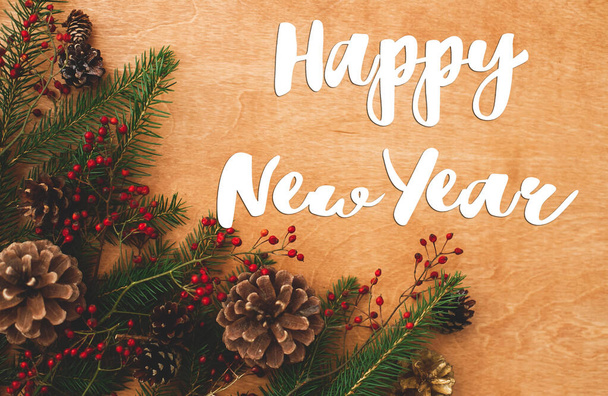 Szczęśliwego Nowego Roku. Szczęśliwego Nowego Roku tekst ręcznie napisany na bożonarodzeniowych gałązkach jodły, czerwone jagody i szyszki sosnowe na drewnie rustykalnym, płaskie układanki. Pozdrowienia, wesołych świąt - Zdjęcie, obraz