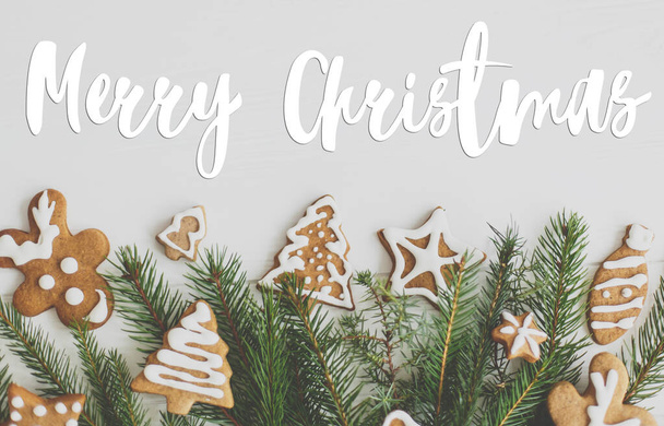 クリスマスのジンジャーブレッドクッキーに手書きのメリークリスマステキストと白い木製の背景フラットレイアウトにモミの枝の境界線。グリーティングカード、現代の季節の挨拶。幸せな休日 - 写真・画像