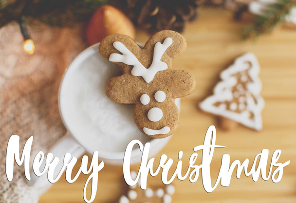 Καλά Χριστούγεννα κείμενο χειρόγραφο σε τάρανδο μπισκότο μελόψωμο για καφέ στο πουλόβερ, κουκουνάρια, κλαδιά ελάτης και φώτα. Ευχετήριες κάρτες, μοντέρνες εποχές. Καλές γιορτές. - Φωτογραφία, εικόνα