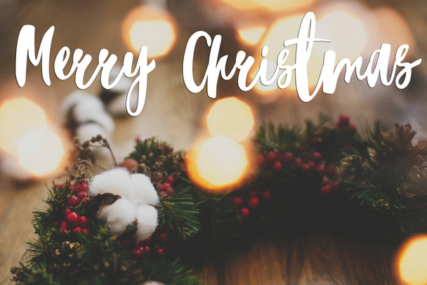 コットン、赤いベリー、ライトの素朴な木の上に松のコーンとクリスマスリースに手書きのメリークリスマステキスト。グリーティングカード季節の挨拶。幸せな休日 - 写真・画像