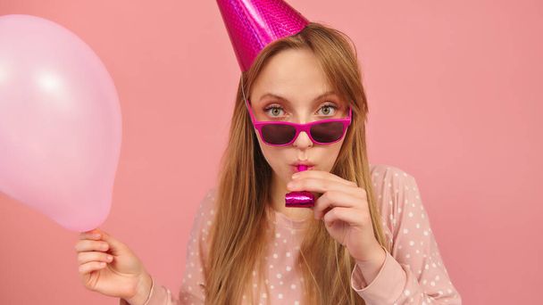 Young party woman blowing party horn whie holding balloon. Isolated on pink background - Photo, Image