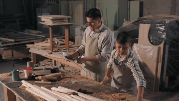 Średni materiał filmowy małego chłopca w koszuli w kratkę i fartuchu zgodnie z instrukcjami ojca zbierającego drewniane szyny na stos na projekt stolarski diy - Materiał filmowy, wideo