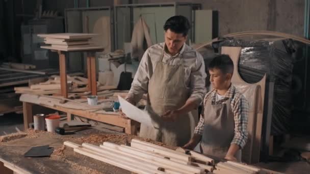 Medium shot of father and son spending time together in carpentry workshop looking at blueprint of diy furniture made of wood - Materiaali, video