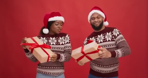 Νεαρό ευτυχισμένο ζευγάρι Αφροαμερικανών σε καπέλα Σάντα κρατώντας χριστουγεννιάτικα δώρα και χορεύοντας πάνω από το κόκκινο φόντο στούντιο - Πλάνα, βίντεο