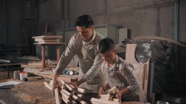 Mediana toma de feliz padre e hijo trabajando en taller de carpintería haciendo cosas de listones de madera, hombre dando instrucciones y niño escuchando cuidadosamente - Metraje, vídeo
