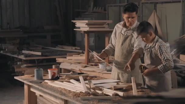 Średnie ujęcie chłopca słuchającego instrukcji ojca, mierzącego kawałek drewna za pomocą trampoliny przy stole ciesielskim w stolarni produkującej diy furniture - Materiał filmowy, wideo