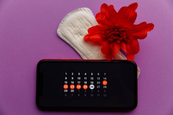 Menstruation app in smartphone with sanitary pad and red flower on a lilac background. Woman critical days and hygiene protection concept - Photo, Image