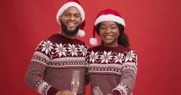 Happy New Year congratulation. Joyful black man and woman in Santa hats tasting to camera with champagne - 映像、動画