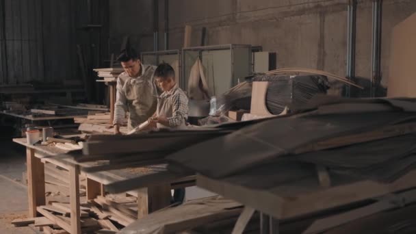 Medium footage of small cluttered carpentry workshop with piles of wood for diy and young boy helping his father working as carpenter to sand wood with sandpaper - Materiaali, video