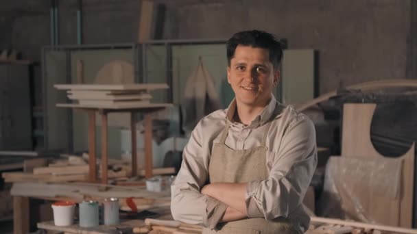 Medio primer plano retrato de artesano en camisa vintage y delantal de pie con las manos dobladas en acogedor taller de carpintería sonriendo a la cámara - Metraje, vídeo