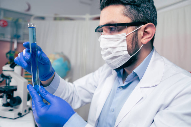 Científico joven mira un tubo de ensayo en el laboratorio. El hombre usa gafas protectoras - Foto, Imagen