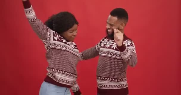 Молода афроамериканська пара в новорічних светрах енергійно танцює, насолоджуючись різдвяною вечіркою, червоним фоном. - Кадри, відео
