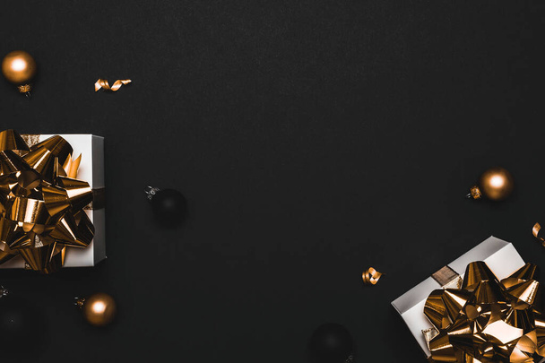 Geschenkboxen Hintergrund. Weiße Geschenkschachtel mit goldenem Band, Neujahrskugeln und funkelnden Lichtergirlanden in weihnachtlicher Komposition auf schwarzer Grußkarte. Winterfestliche Komposition mit Kopierraum - Foto, Bild