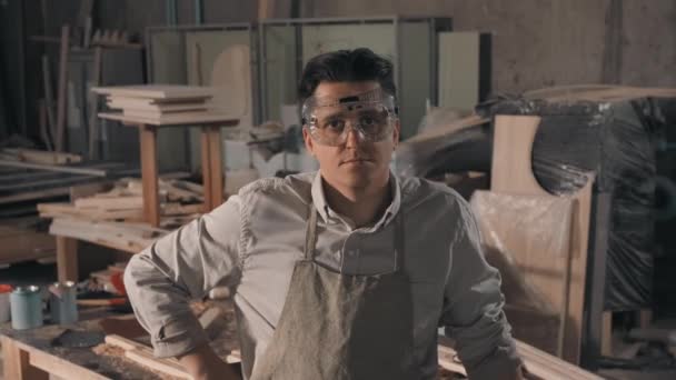 Medium close-up portrait of man working as carpenter in small joinery looking at camera wearing safety glasses for woodworking with piles of wood planks in background - Filmati, video