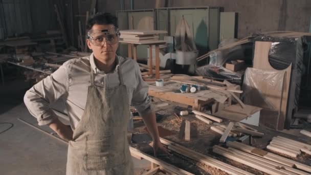 Medium portrait of caucasian adult man in safety glasses wearing vintage shirt and carpenter apron looking at camera with cluttered carpentry workshop for woodworking in background - 映像、動画