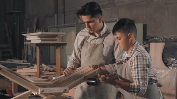 Medium shot of young boy studying carpentry art helping father nailing wooden chair with hammer in small cozy with piles of wooden planks - Filmagem, Vídeo