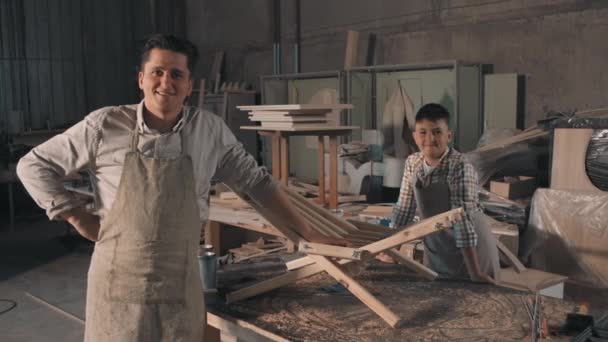 Mittleres Porträt von zwei fröhlichen Handwerkern in Schürzen, die gerade gemeinsam in einer kleinen Tischlerei Holzdiy-Liegestühle gefertigt haben - Filmmaterial, Video