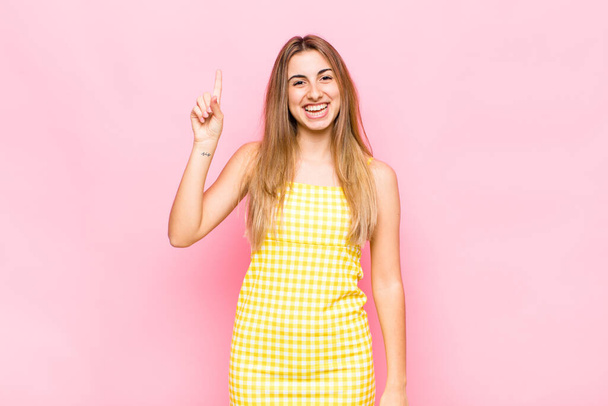 blonde woman smiling cheerfully and happily, pointing upwards with one hand to copy space - Foto, afbeelding