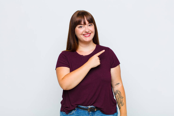 overweight woman smiling cheerfully, feeling happy and pointing to the side and upwards, showing object in copy space - Foto, immagini