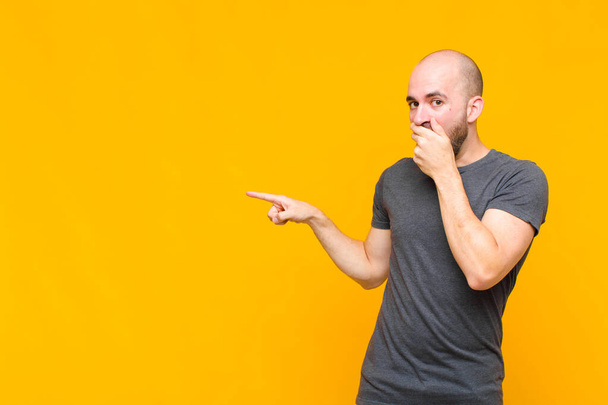 bald man feeling happy, shocked and surprised, covering mouth with hand and pointing to lateral copy space - Foto, Imagen
