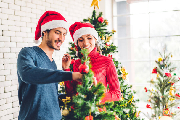 Ρομαντικό γλυκό ζευγάρι σε Σάντα καπέλα διασκεδάζοντας διακόσμηση χριστουγεννιάτικο δέντρο και χαμογελώντας ενώ γιορτάζει την παραμονή της Πρωτοχρονιάς και απολαμβάνοντας το χρόνο που περνούν μαζί τα Χριστούγεννα στο σπίτι - Φωτογραφία, εικόνα