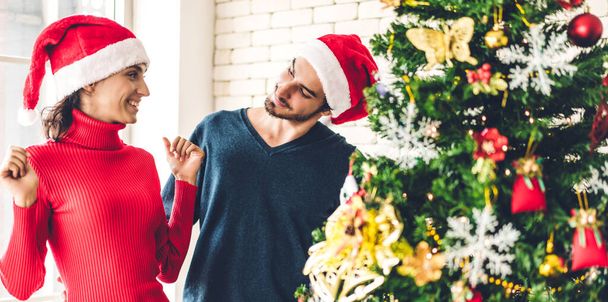 Noel Baba şapkalı romantik çift Noel ağacını süsleyerek ve gülerek Noel arifesini kutlarken ve Noel zamanı evde birlikte vakit geçirirken eğleniyor. - Fotoğraf, Görsel