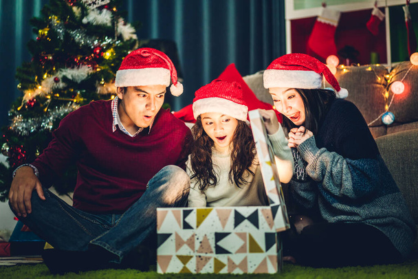 Porträt eines glücklichen Familienvaters und einer Mutter mit Tochter in Weihnachtsmützen, die Spaß daran haben, eine magische Weihnachtsgeschenkbox zu öffnen und die gemeinsame Weihnachtszeit zu Hause zu genießen - Foto, Bild