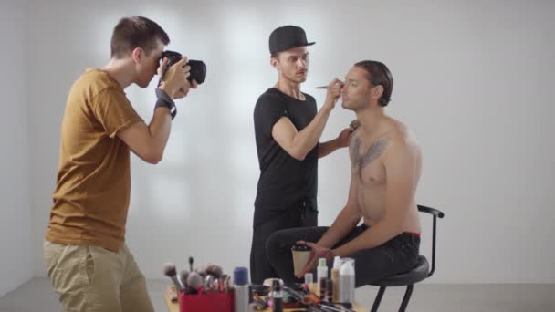 Середній знімок фотографа, який фотографує чоловіка-косметолога, який робить макіяж на безсорочному чоловічому клієнті, що сидить на стільці в студії
 - Кадри, відео